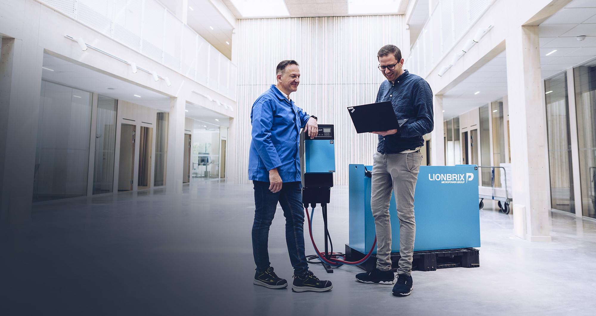 Two male employees at Micropower Group are standing beside a battery charger talking and looking in a laptop.