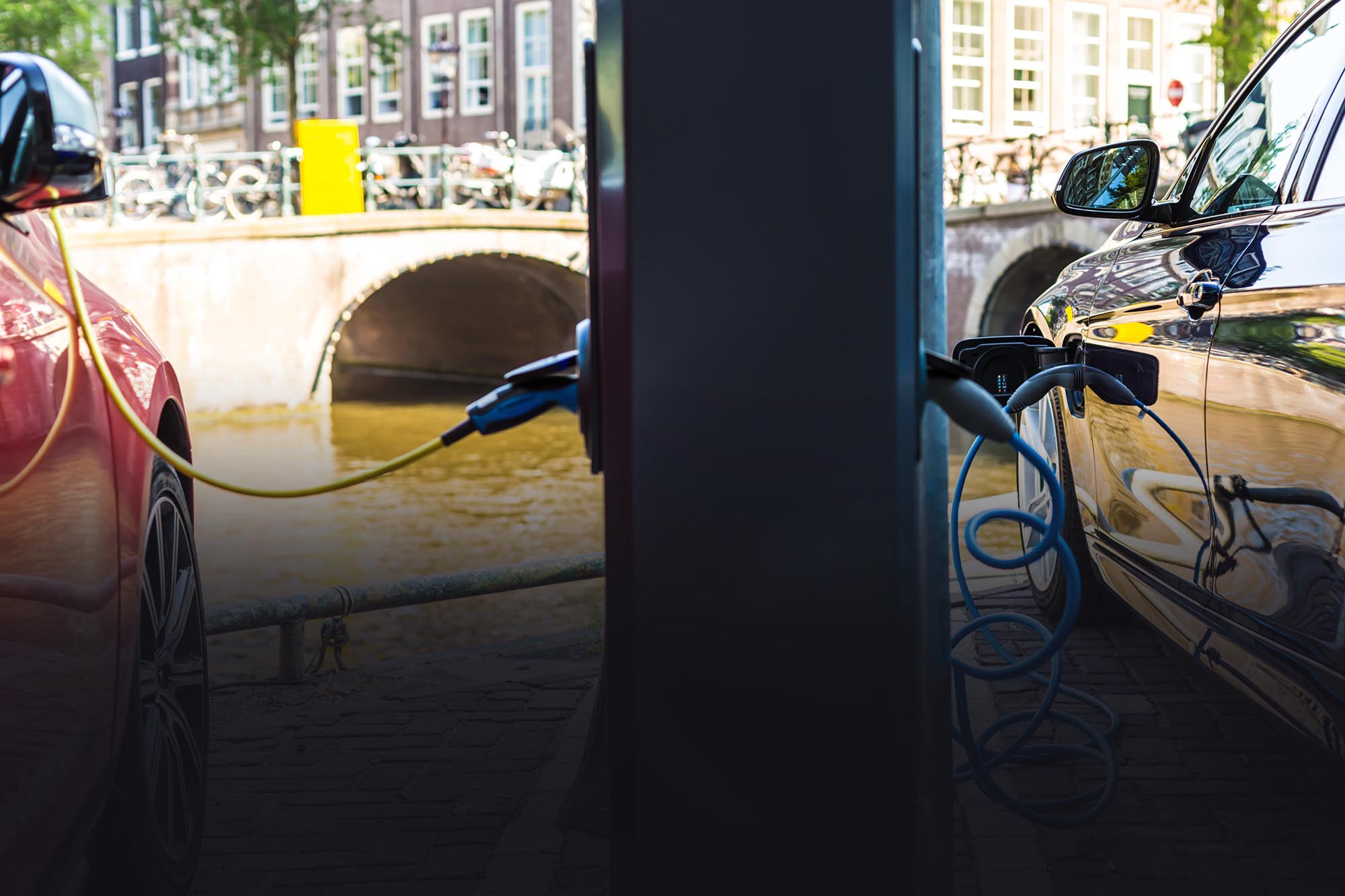 Two modern electric cars, one red and one yellow, is being charged in the city, by a river.