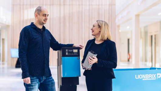 A man and a woman is talking, standing on each side of a blue industrial battery charger.